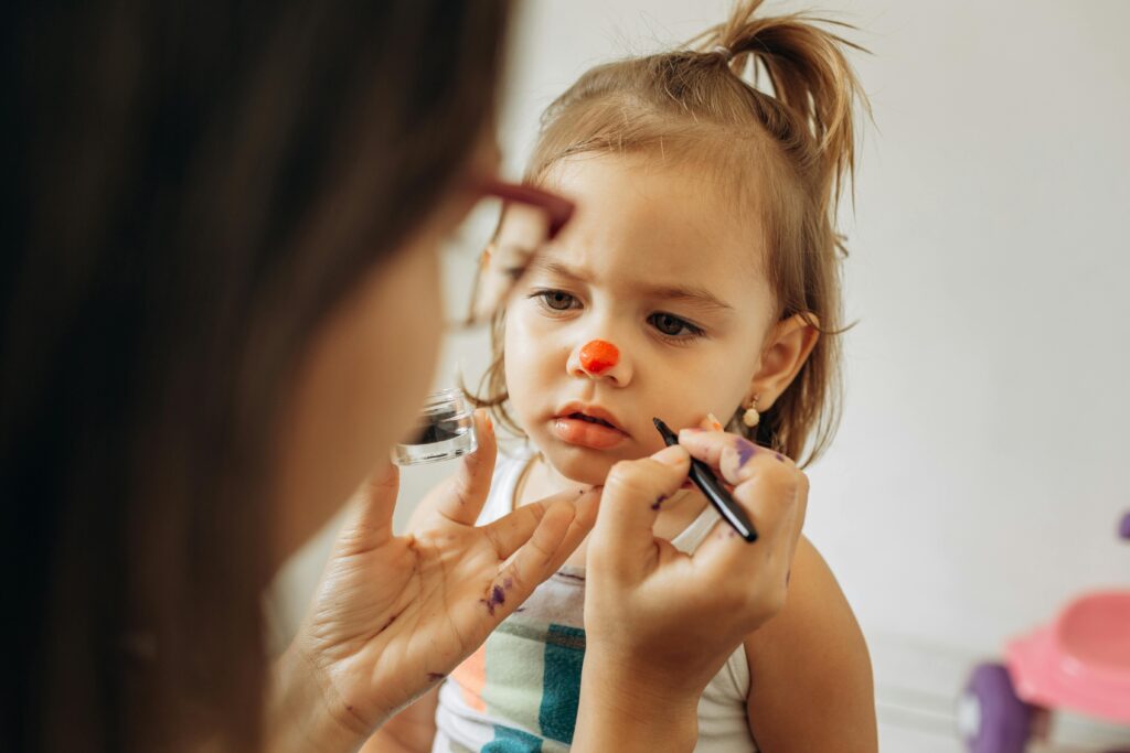 A little girl gets her face painted by a woman in a playful and creative indoor setting.